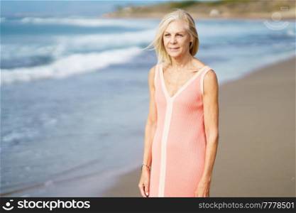 Serene senior female in dress and with gray hair standing on sandy coast near sea in summer and looking away. Peaceful elderly woman on beach in summer