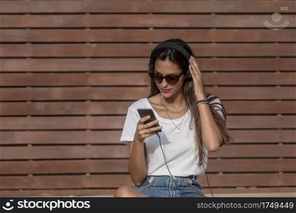 Serene woman in white shirt with red lipstick relaxing on wooden bench while listening to music in headphones and using smart phone in summer. Pensive female in earphones chilling in park