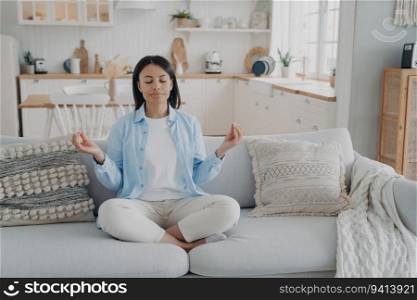 Serene woman practices yoga, meditates on couch in lotus position. Breathing exercises for mental balance, anxiety relief. Healthy lifestyle at home.
