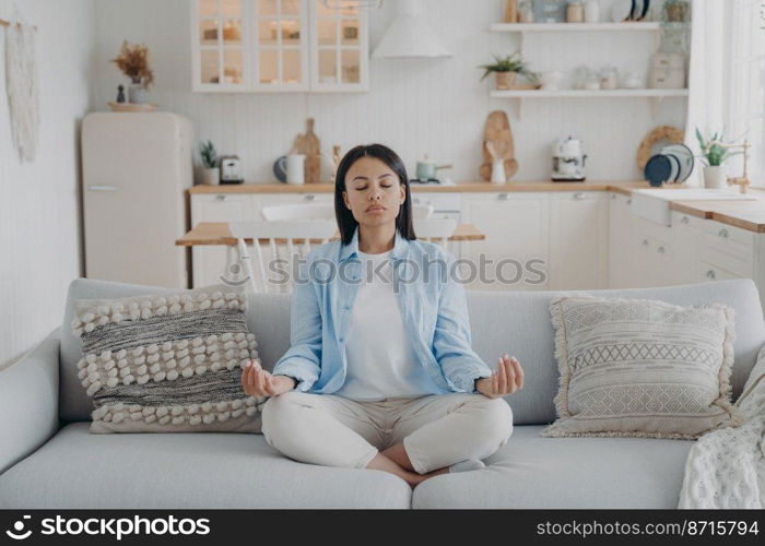 Serene woman practicing yoga and meditation keeping hands in mudra gesture, sitting cozy couch at home. Calm young female relaxing on sofa. Wellness, stress relief, emotion management.. Serene woman practices yoga meditation sitting on cozy couch at home. Wellness, stress relief