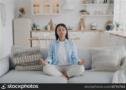 Serene woman practicing yoga and meditation keeping hands in mudra gesture, sitting cozy couch at home. Calm young female relaxing on sofa. Wellness, stress relief, emotion management.. Serene woman practices yoga meditation sitting on cozy couch at home. Wellness, stress relief