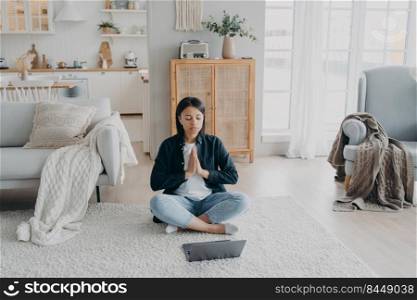 Serene young woman practice yoga at laptop while online lesson sitting on floor in living room. Calm female meditates having break after working on computer at home. Stress relief, healthy lifestyle.. Calm woman practices yoga at laptop, has break after working sitting on floor at home. Stress relief