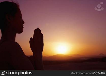 Serene young woman with hands together in prayer pose in the desert in China, silhouette, sun setting, profile