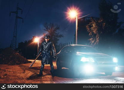 Serial maniac in leather coat and hat with bloody baseball bat wrapped in metal chain against black car with light at the night. Horror, bloody murderer, murder weapon