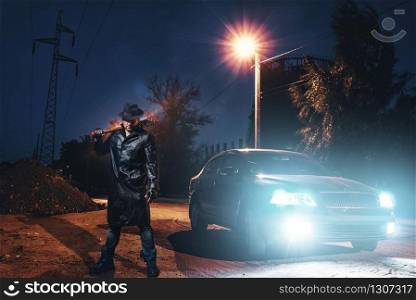 Serial maniac in leather coat and hat with bloody baseball bat wrapped in metal chain against black car with light at the night. Horror, bloody murderer, murder weapon. Maniac with bloody baseball bat against black car