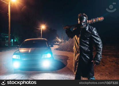 Serial maniac in leather coat and hat with bloody baseball bat wrapped in metal chain against black car with light at the night. Horror, bloody murderer, murder weapon. Maniac with bloody baseball bat against black car