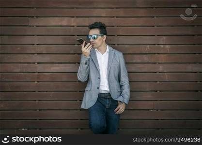 Serious adult male in sunglasses wearing stylish jacket and jeans leaning against wooden wall and speaking on mobile phone while using voice recognition. Trendy businessman using voice recognition on smartphone