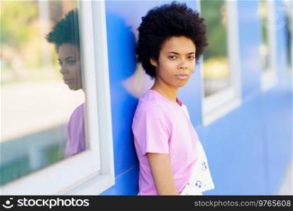 Serious African American female with Afro hairstyle looking away with thoughtful face while standing near blue building with windows on street of city. Black woman standing near blue house