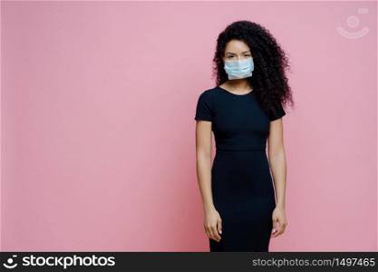 Serious Afro American woman wears disposable medical mask on face, being on self isolation during quarantine, stays at home alone, has symptoms of coronavirus, stands against pink background
