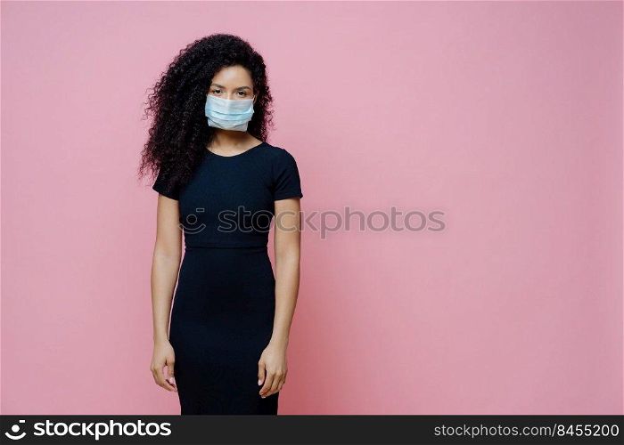 Serious Afro American woman wears disposable medical mask on face, being on self isolation during quarantine, stays at home alone, has symptoms of coronavirus, stands against pink background