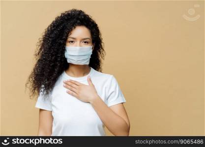 Serious Afro American woman wears medical face mask, has problems with breathing, presses hand to chest, got infected with coronavirus, isolated on beige ackground. Covid 19, health care concept