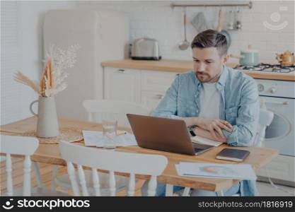 Serious and focused young freelancer working on laptop while sitting by kitchen table with hands resting on it, graphs and statistics scattered around, open note book next to him. Focused young freelancer working on laptop while sitting by kitchen table