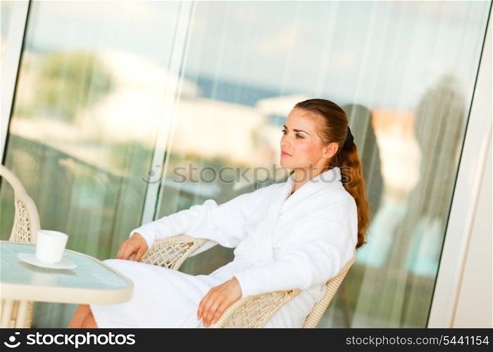 Serious attractive female in bathrobe sitting at table on terrace with cup of coffee&#xA;