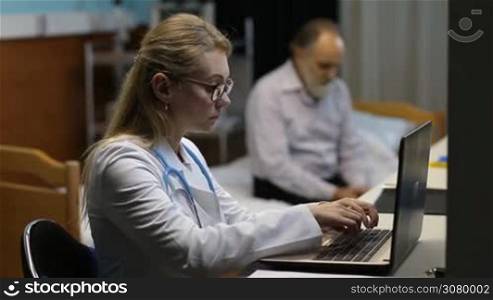 Serious attractive female physician in eyeglasses and lab coat working with laptop in medical office with worried senior ill patient on background. Confident woman doctor writing and filling out a prescription in front of laptop pc.
