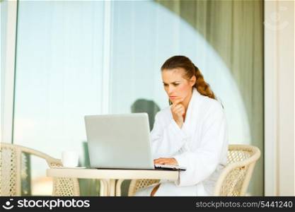 Serious attractive woman in bathrobe sitting at table on terrace and working on laptop&#xA;