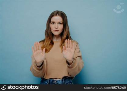 Serious attractive young woman with brown hair in casual wear holding hands in front of her demonstrating stop gesture while posing on blue light blue studio background. Pretty female demonstrating stop gesture while posing against blue wall