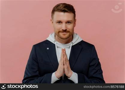 Serious bearded adult man keeps palms pressed together prays for something looks directly at camera believes in something good happen poses against pink background. Handsome guy makes request