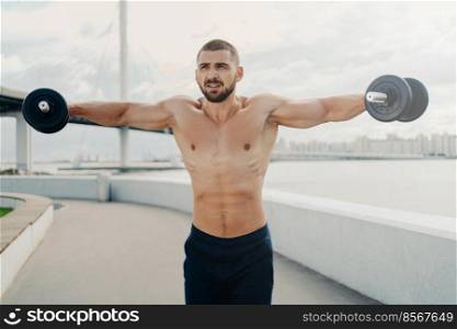 Serious bearded athlete man raises barbells puts all efforts in gaining strong muscles on arms has well built body poses outdoor near river. Determined sportsman has weightlifting exercise outside