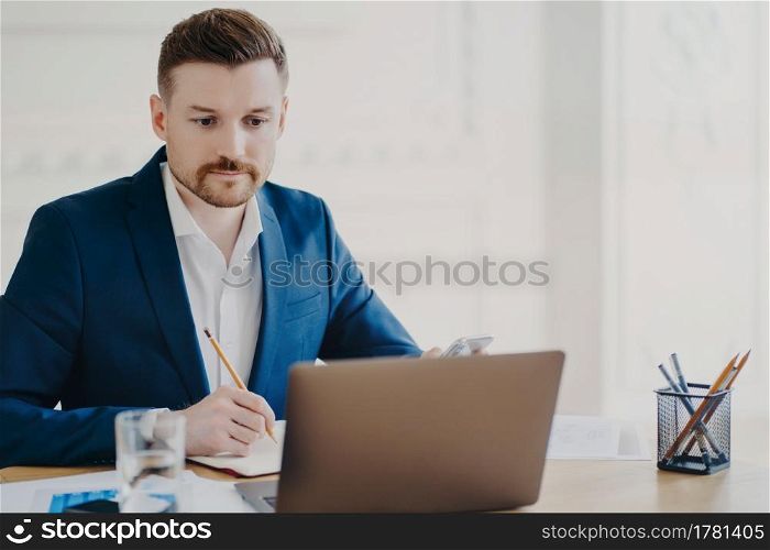 Serious bearded man marketer involved in working process makes notes with pencil looks attentively at laptop computer wears blue formal suit poses at coworking space writes organisation plan