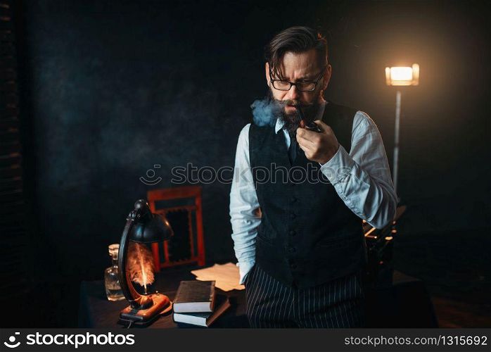 Serious bearded writer in glasses smoking a pipe. Feather, crystal decanter, books and vintage lamp on the desk. Serious bearded writer in glasses smoking a pipe