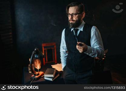 Serious bearded writer in glasses smoking a pipe. Feather, crystal decanter, books and vintage lamp on the desk. Serious bearded writer in glasses smoking a pipe