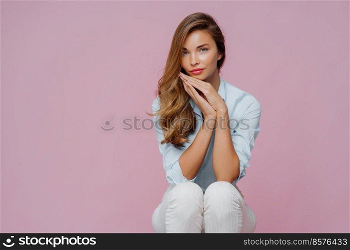 Serious beautiful woman with long hair, dressed in shirt and trousers, keeps palms pressed together undr chin, has confident calm look, sits on chair over violet background, copy space area aside