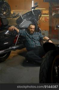 Serious biker sitting on floor and thinking searching way to repair motorcycle. Motorbike garage service and hobby concept. Serious biker sitting on floor and thinking searching way to repair motorcycle