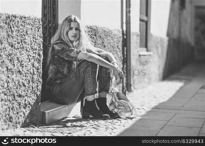 Serious blond woman, model of fashion, sitting on floor in urban background. Beautiful girl wearing shirt and blue jeans.