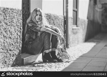 Serious blond woman, model of fashion, sitting on floor in urban background. Beautiful girl wearing shirt and blue jeans.
