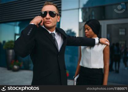 Serious bodyguard in suit and sunglasses requests support on earpiece for female client protection. Security guard is a risky profession, professional guarding