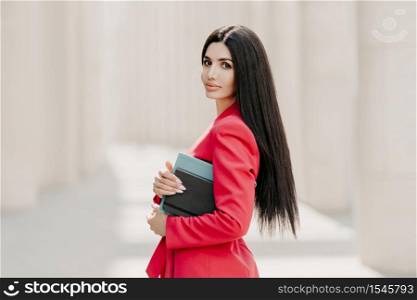 Serious brunette businesswoman with makeup, manicure, wears red jacket, carries diary and notepad, stands in profile outdoor, goes for work. Elegant businesslady on street. Stylish director outside