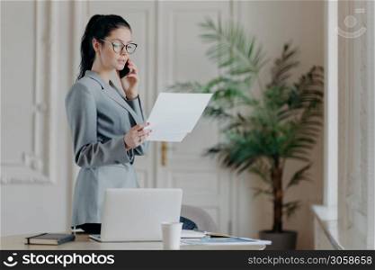 Serious brunette female employee stands near desktop, holds paper documents, talks via smartphone, wears formal clothes and spectacles, discusses business strategy with partner, organizes meeting