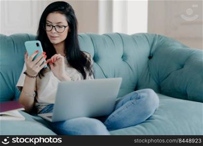 Serious brunette female freelancer works remotely on laptop computer, sends text messages via smartphone connected to wireless free internet sits crossed legs on comfortable sofa surfs social networks