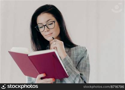 Serious brunette female management consultant uses notepad for planning organization, holds pen and writes down notes or list to do, wears formal clothing, has serious expression, looks in notebook