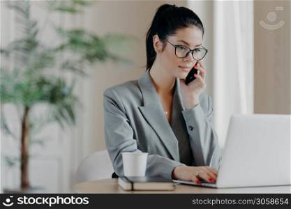 Serious brunette woman keyboards information, concentrated into laptop computer, dressed in formal outfit, poses at coworking space, works remotely, involved in working process during daytime.