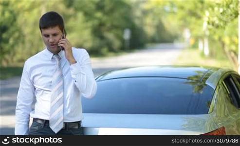 Serious business executive in formal wear talking on smartphone with his business partner while staniding in front of his parked car on roadside. Busy businessman communicating on mobile phone outdoors.