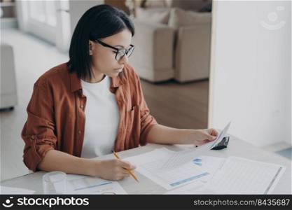 Serious business lady is examining documents. Young caucasian woman working remote from home. Manager or entrepreneur doing paperwork. Concept of intelligent professional and business strategy.. Serious business lady is examining documents. Intelligent professional and business strategy.