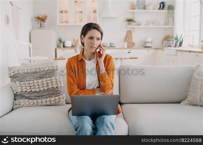 Serious businesswoman talking on mobile phone, sitting with laptop on sofa at home. Frowning female arguing by mobile, solving problem, dissatisfied with bad customer support during phone negotiations. Female arguing by phone, dissatisfied with bad customer support, sitting with laptop on sofa at home