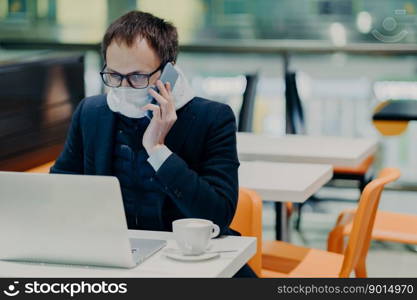 Serious busy man worker works in quarantine, reads information online on laptop computer, wears protective medical mask, calls someone via cellular, cares about health during epidemic virus infection