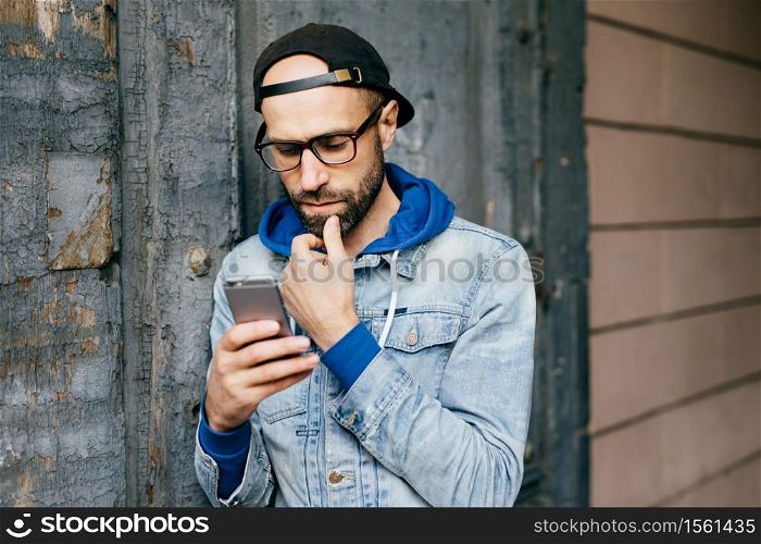 Serious concentrated bearded man in cap and denim jacket standing against cracked wall holding smartphone liking posts and sharing them via social networks, reading news with deep and thoughtful look