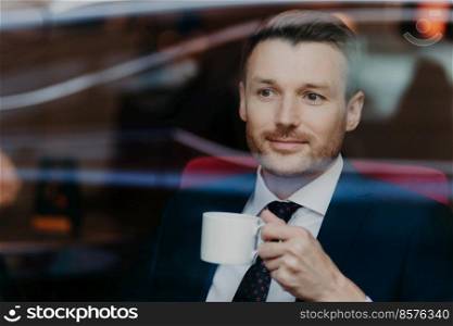 Serious concentrated unshaven male entrepreneur looks thoughfully in window of coffee shop, drinks tea, thinks about future meeting with partners, wears elegant clothing. Coffee break concept