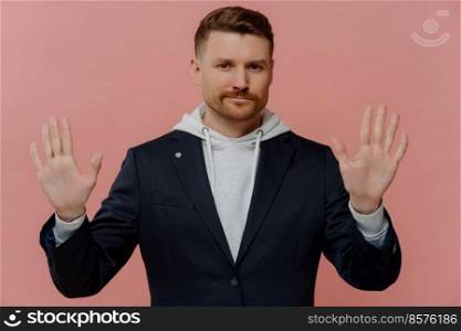 Serious confident adult man with bristle raises hands towards camera shows palms has strict expression asks to hold on or stop wears comfortable hoodie and jacket isolated over pink background