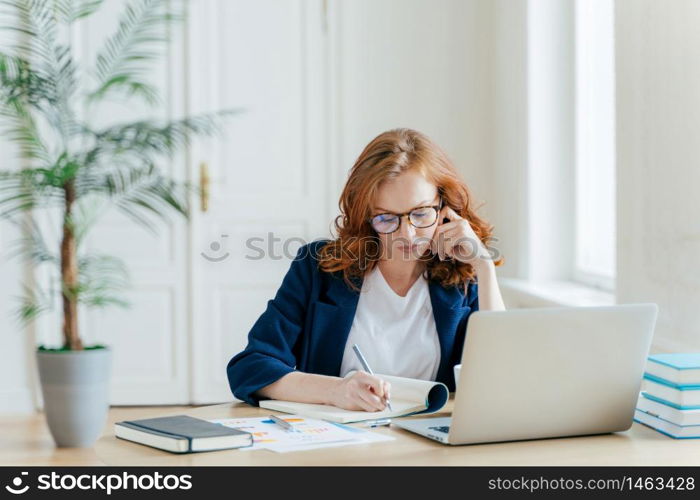 Serious confident female entrepreneur in formal clothes writes down information while watches business webinar, notes main ideas, sits at work place in front of opened computer makes notes for startup