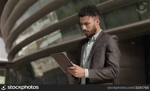 Serious confident man wearing formal wear holding tablet. Handsome young businessman with tablet standing near modern building. Business and technology concept