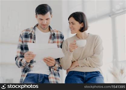 Serious couple study documents together, have serious looks, drink coffee, dressed in casual wear, plan their budget, pose in spacious light room, do paperwork, busy preparing financial report