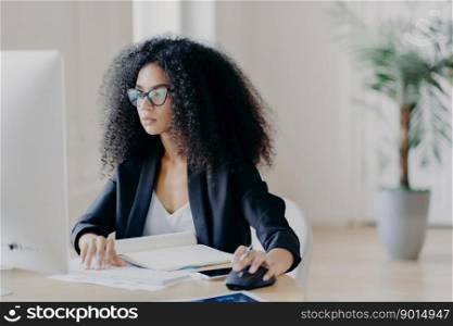 Serious curly businesswoman focused at display of computer, works on making project, surrounded with textbook and papers, wears glasses for vision correction, black suit, poses in office room