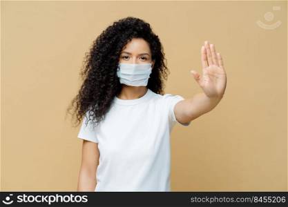 Serious dark skinned woman makes stop gesture, pulls palm towards camera, wears medical flu mask, asks stay at home not to spread coronavirus disease, prevents virus, dressed in white t shirt.