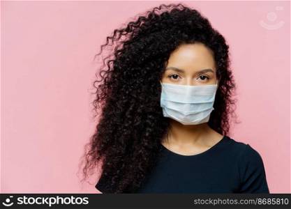 Serious dark skinned young woman being on self isolation at home, wears protective medical mask, being on quarantine at home, dressed casually, poses against pink background. Coronavirus disease