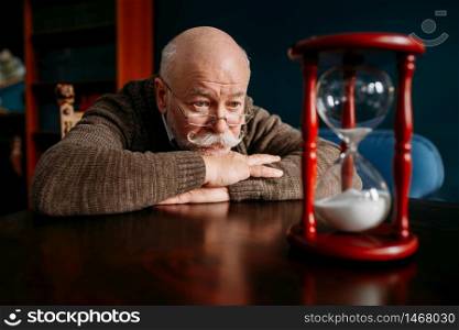 Serious elderly man looking on sandglass in home office, time cannot be turned back. Mature senior looking on hourglass. Elderly man looking on sandglass in home office