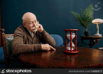 Serious elderly man looking on sandglass in home office, time cannot be turned back. Mature senior looking on hourglass. Elderly man looking on sandglass in home office